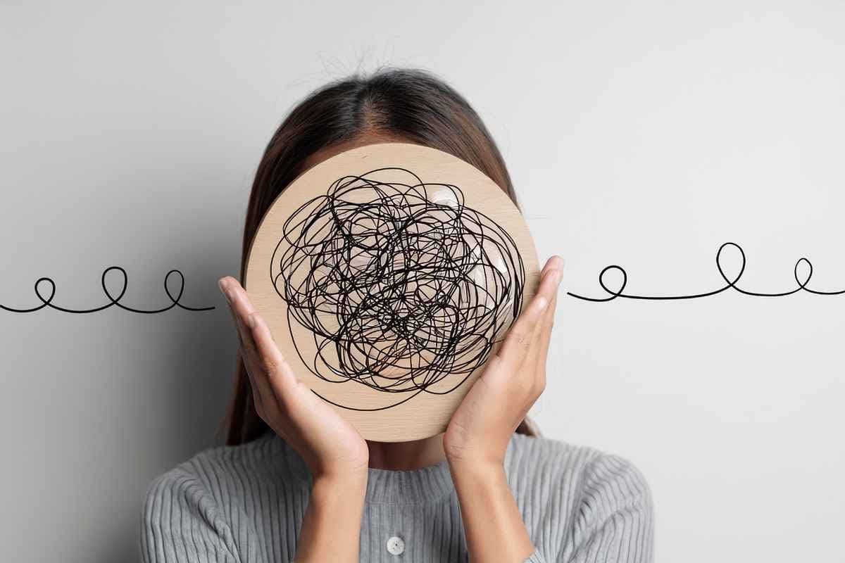 A woman holds a wooden disc covered in tangled scribbles in front of her face, a tangle of thoughts hiding who she is.