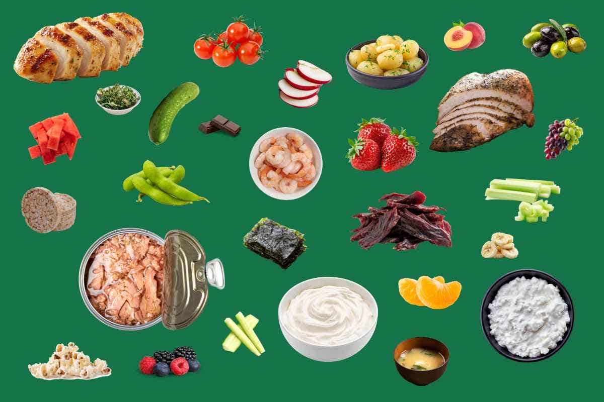 A spread of low calorie snacks on a green background: chicken breast, prawns, tinned tuna, cottage cheese, boiled new potatoes, strawberries, edamame and vegetable sticks.