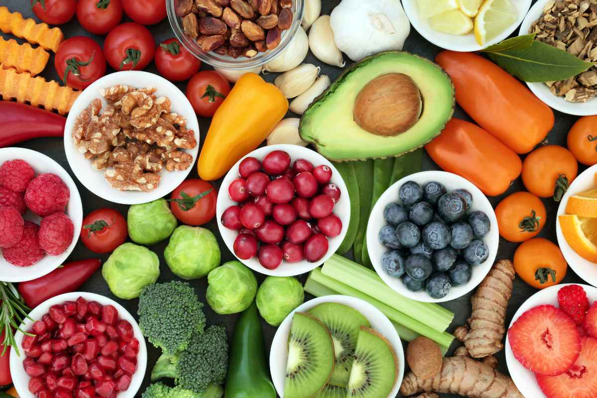 An overhead spread of fresh vegetables, fruit, berries and nuts in small white bowls.