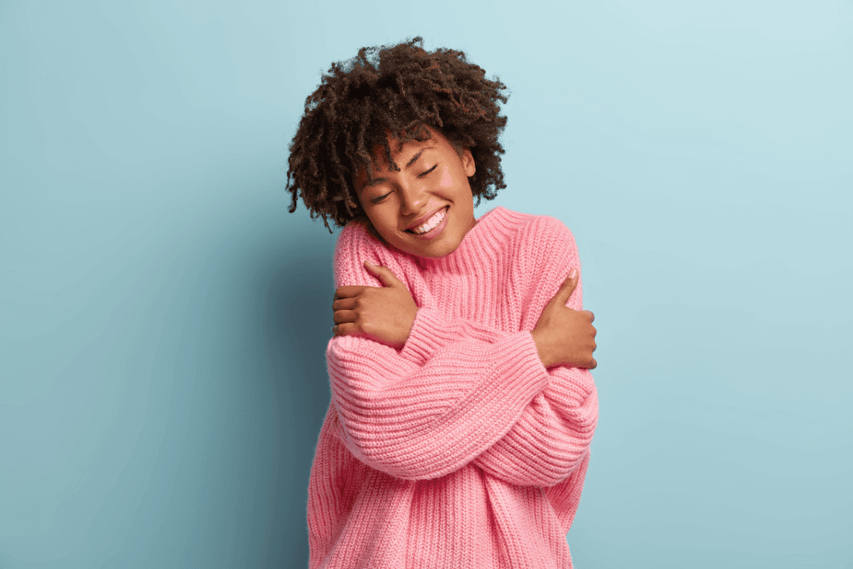 A smiling person with curly hair, in a loose pink knitted sweater, hugs themselves with their eyes closed against a pale blue wall.