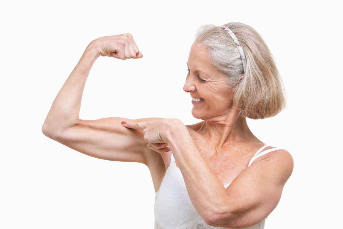 An older woman flexing her bicep and pointing at it, smiling.
