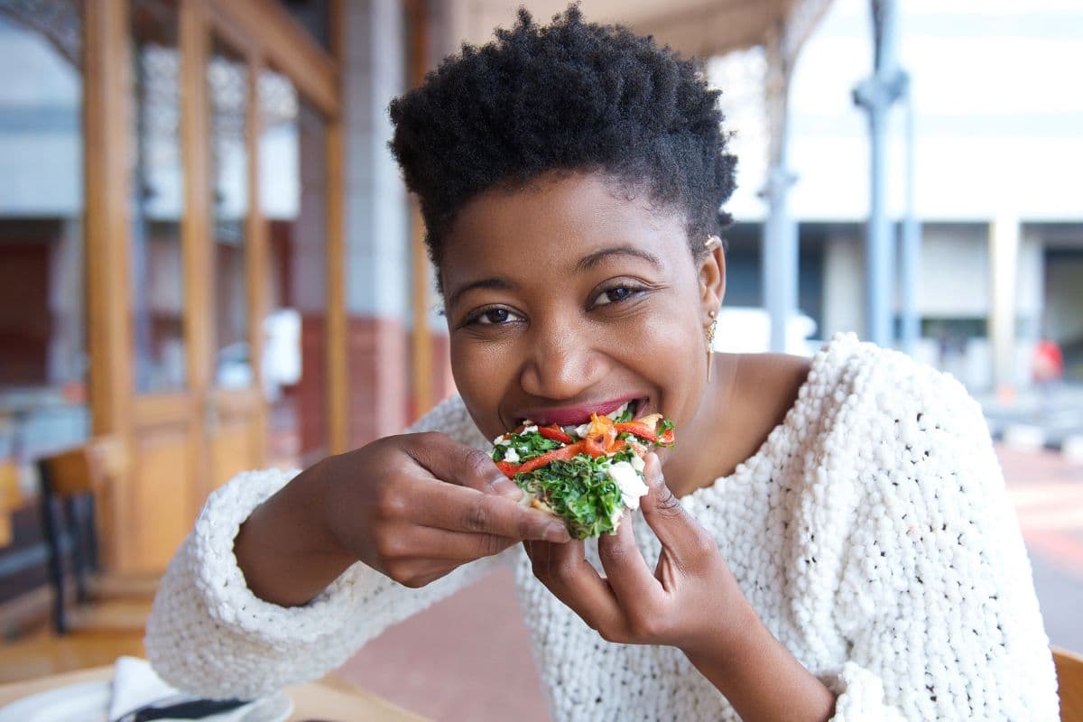 A woman in a chunky white knitted sweater sits at an outdoor cafe table and smiles as she bites into a sandwich filled with greens, red pepper and soft cheese.